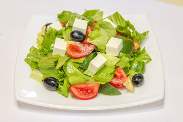 Photo of a Greek salad on a white plate with tomatoes, olives, feta cheese and herbs.