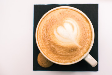 Italian cappuccino mug flat lay background. Black coffee with white blossoming flower or heart shape image on milk froth with small biscuit on decorative square black paper tissue on white table top.