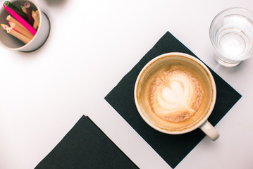 Creative minds morning coffee or tea, glass of water and colourful pencils on white table top flatlay background. Half drunk Italian cappuccino mug on black serviettes, flower or heart on milk froth.
