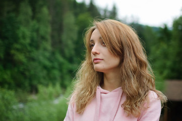 Attractive young calm blonde woman in pink clothes with long hair on the hill with green forest background during spring or early autumn in the mountains.