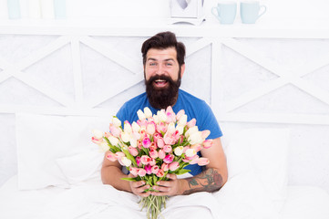 Spring mood in his bedroom. Man hold tulips bouquet while relaxing in bed. Flowers delivery service. Birthday anniversary holiday. Fresh flowers surprise. Gift for spouse. Bearded hipster in bed