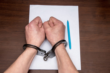 A man with bare hands in handcuffs sits at a table in front of a blank sheet of paper and a fountain pen. Concept: the detainee writes a confession, the prisoner signs the document.