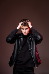 Obraz premium Attractive young guy posing in studio. Young caucasian guy model in black clothes posing in the studio on a dark burgundy background with red backlight. Man in a leather jacket