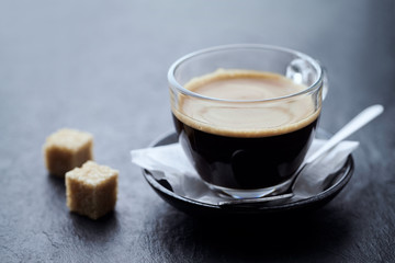  Coffee in glass cup and two brown sugar cubes on dark stone background. Close up.