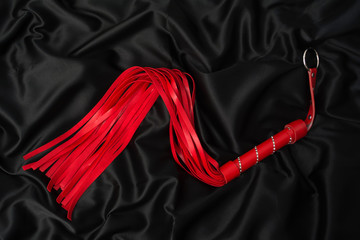 Red whip on a black silk background. Accessories for adult sexual games - obrazy, fototapety, plakaty