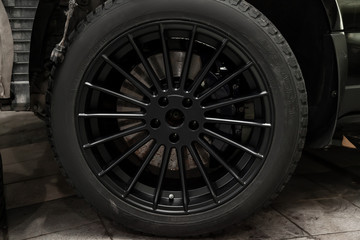 An aluminum alloy wheel with winter studded tires on a black car with a new brake disk requiring replacement. Weather and transport. Auto service industry.