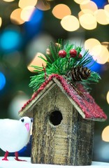 Toy bird standing next to a decorated bird house ornament with Christmas tree lights blurred in the background. Vertical image can be used as a holiday card.