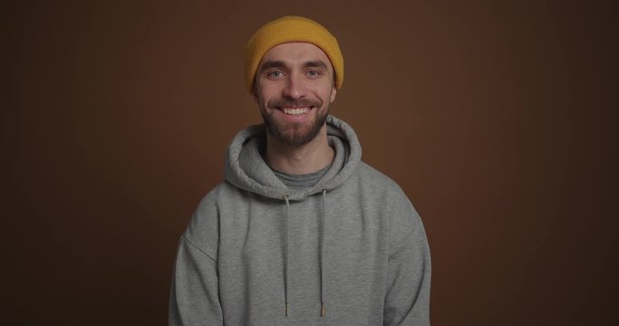 Portrait of guy in gray hoodie and yellow hat smile to camera brown background