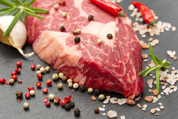 Raw rib eye steak with spices and vegetables. Ingredients for restaurant meal. Fresh meat, salt, rosemary, thyme, chilli, cherry tomatoes, garlic on black stone. Food background.