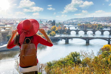 A traveler woman with red sunhat sits on a hill above Prague, Czech Republic, and enjoys the view to the city on a sunny day - obrazy, fototapety, plakaty