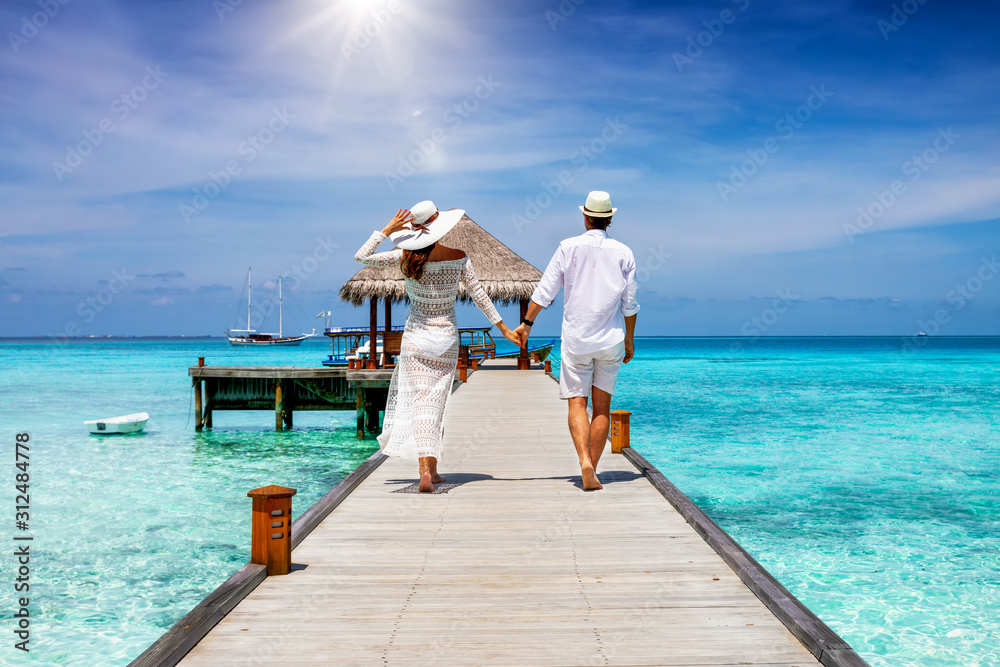 Wall mural a happy couple in white summer clothing on vacation walks along a wooden pier over tropical, turquoi - Wall murals
