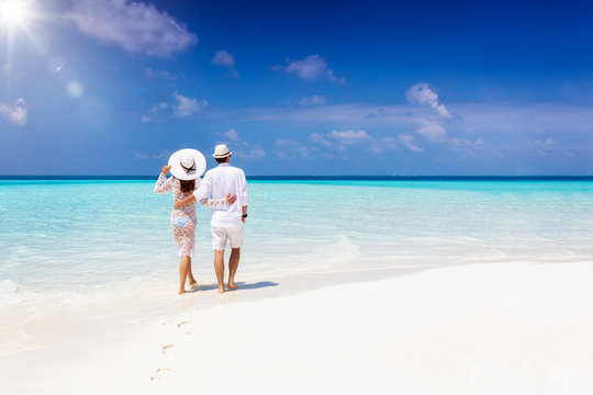 A hugging honeymoon couple walks down a tropical beach with turquoise sea and sunshine 