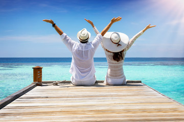 Glückliches Paar im tropischen Urlaub sitzt auf einem Holzsteg über türkisem, Ozean und genießt...