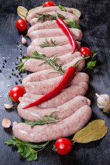 Uncooked pork sausages among vegetables, greens, spices on dark surface