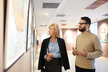 Waist up portrait of mature couple looking at paintings while enjoying exhibition in art gallery or...