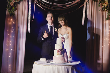 bride and groom stand next to a large wedding cake outside. wedding day