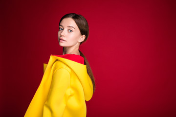 Side view of calm peaceful young woman posing on camera. Wear yellow coat with hood. Stand alone and pose on modern clothes. Isolated over red background.