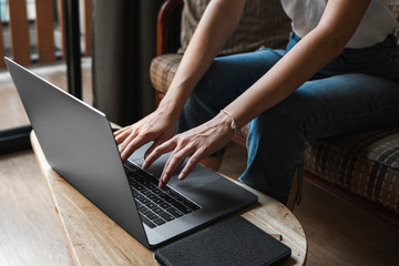 Fototapeta premium Woman is sitting on a sofa and using a laptop at wooden table. Study and work online, freelance. Self employed girl is working with her notebook sitting on a couch with a phone and ereader on table.