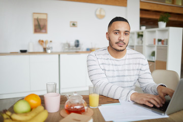 Fototapeta na wymiar Portrait of handsome mixed-race man looking at camera while working from home and using laptop at kitchen table, copy space