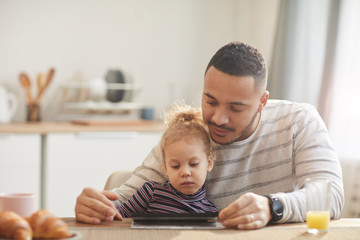 Warm-toned portrait of caring father with cute little girl using digital tablet together while sitting at wooden table in cozy kitchen, copy space