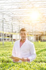 Portrait of confident scientist standing with clipboard amidst herbs in greenhouse
