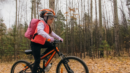 Obraz premium One caucasian children rides bike road in autumn park. Little girl riding black orange cycle in forest. Kid goes do bicycle sports. Biker motion ride with backpack and helmet. Mountain bike hardtail.