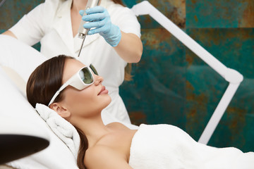beautiful woman receiving facial treatment with laser in beauty salon by beautician