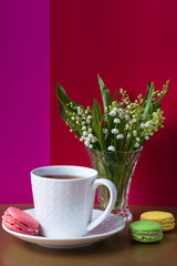 French multicolored cakes macarons and a cup of tea. A bouquet of lily of the valley in a crystal vase.