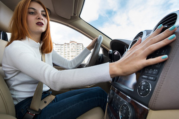 Wide angle view of young redhead woman driver fastened by seatbelt driving a car adjusting heater.