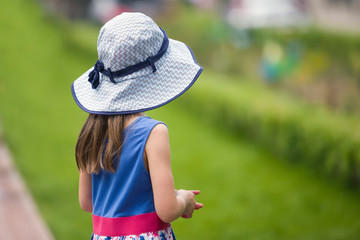 Little child girl in summer dress and a hat walking alone in green park.