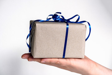 the girl holds in her hand a box in beige Kraft paper with a blue ribbon for a gift on a white background. congratulations on holidays in the color of the year 2020. gift in a hand