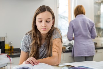 Girl studying at table with mother standing in background