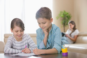 Siblings drawing together at table with mother in background