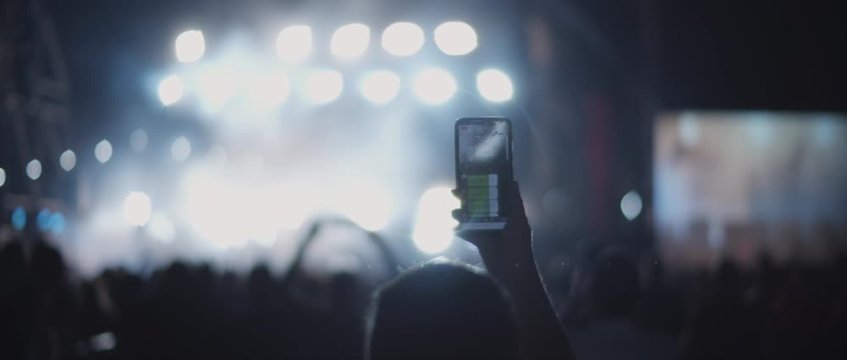 Person taking a picture during a concert, with floodlights flashing in the background. People in front of a stage of a live show at a night club. Medium, behind shot, handheld, slow motion. BMPCC 4K 