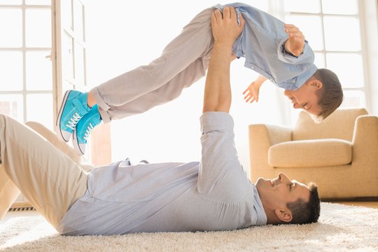 Side View Of Playful Father Lifting Son While Lying On Floor At Home