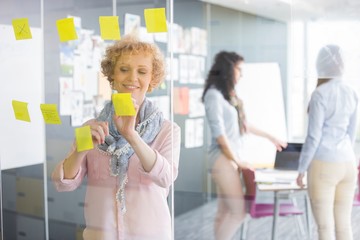 Businesswoman reading sticky notes on glass with colleagues in background
