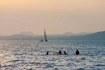 Lake Balaton at sunset in back light with sailboats and silhouettes of bathers.