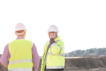 Male engineer using mobile phone while standing with colleague at construction site against clear sky