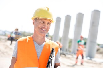 Happy architect looking away while holding clipboard at construction site