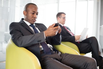 Young businessman using mobile phone on chair with male colleague in background at office