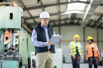 Mature male inspector writing on clipboard while workers in background at industry