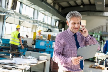 Mature male supervisor looking at clipboard while talking on mobile phone in industry