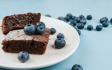  A piece of brownie on a white plate, sprinkled with icing sugar and decorated with blueberries on a blue wooden background copy space. Berry chocolate cake decor.