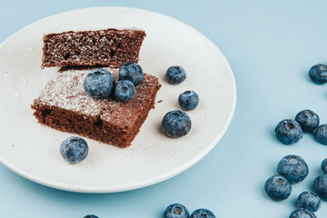  A piece of brownie on a white plate, sprinkled with icing sugar and decorated with blueberries on a blue wooden background copy space. Berry chocolate cake decor.