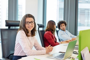 Portrait of smiling young businesswoman with laptop while colleagues in background at office