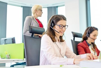 Female customer service representative using headset while colleagues in background at office