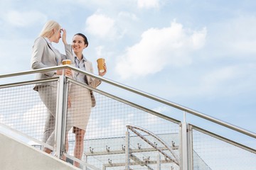 Young businesswomen with disposable coffee cups standing by railing against sky