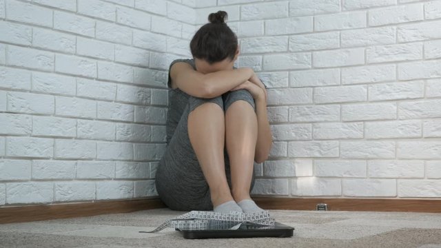 Woman with figure problem. A woman is crying while sitting in the corner in front of a bathroom scale and a measuring tape.