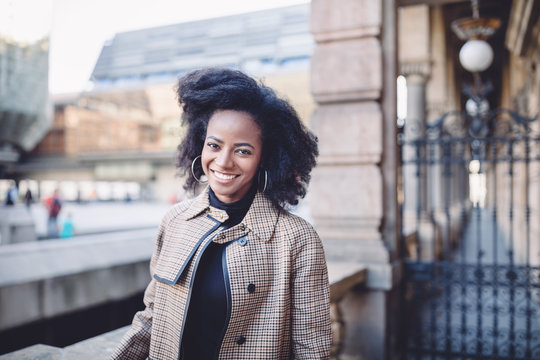 Beautiful african american young woman with afro and large hoop earrings in a stylish coat, smiling. Urban street portrait.