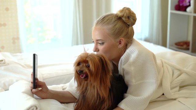 Slow motion of the blonde girl is playing with a dog in the bed Take a selfie with happiness in pajamas. Smiling woman with pet dog yorkshire terrier lying on bed in sleepwear.